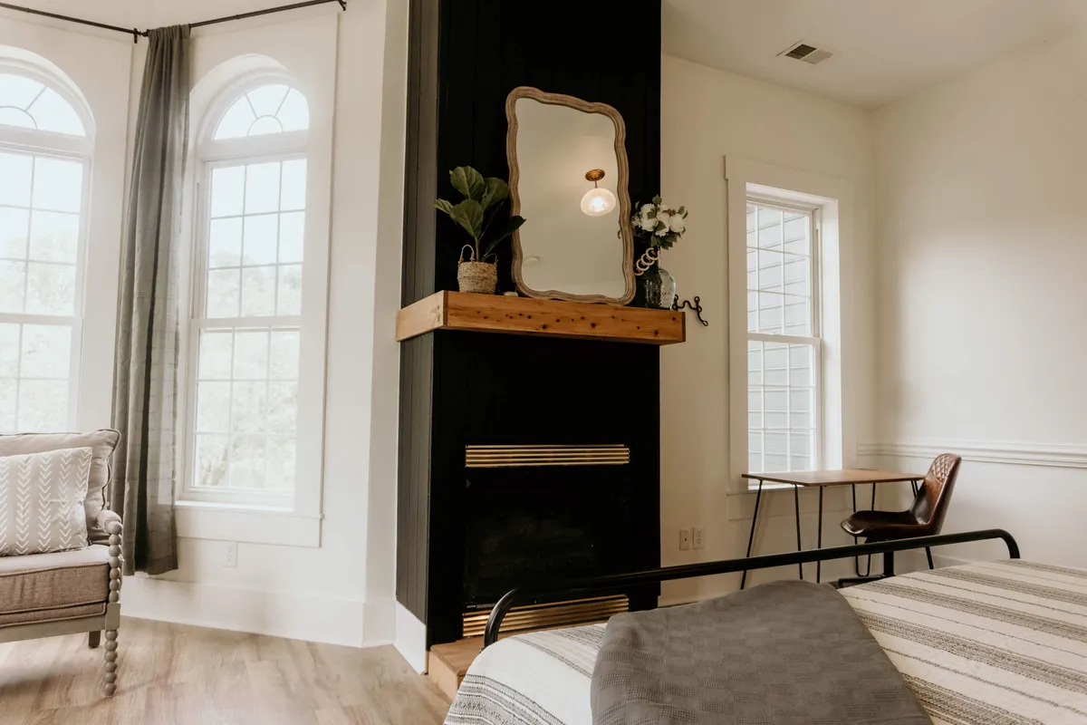 A serene living room scene featuring a classic fireplace flanked by tall windows, an ornate mirror above the mantel, and plush seating arranged for comfort. Gentle sunlight streams in, complementing the soft, warm lighting and inviting atmosphere, while period details such as woodwork and molding enhance the Victorian character.
