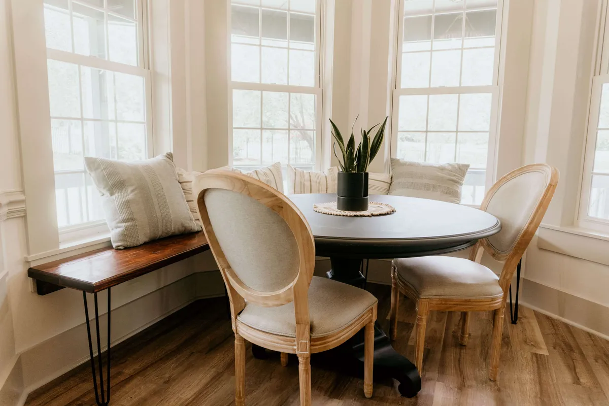 A rounded dining area set beside expansive windows, allowing abundant natural light to flood the space. The table is surrounded by inviting seating and accentuated by potted greenery, making it an ideal spot for morning coffee, quiet reading, or lively conversation.