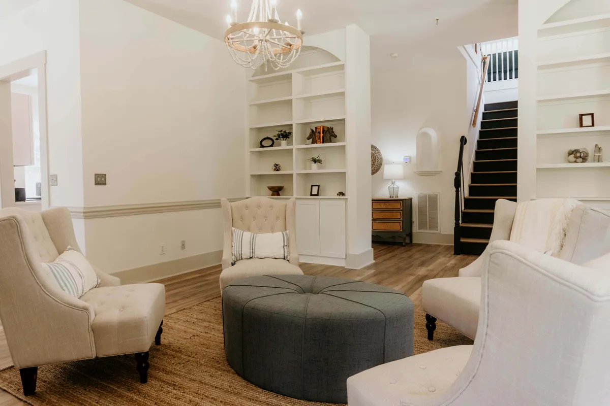A welcoming sitting area highlighted by a sparkling chandelier, built-in shelves filled with books and decor, and comfortable armchairs clustered around a round ottoman. The arrangement encourages conversation, with soft lighting and charming accents enhancing the inviting mood.
