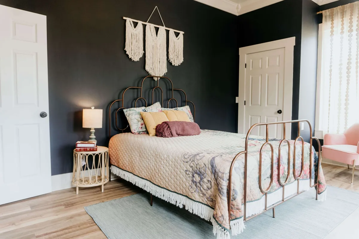 A bedroom featuring a bed with an ornate Victorian-style headboard, vibrant throw pillows, a macrame wall hanging, and a side table illuminated by a lamp. The scene exudes warmth, with natural sunlight filtering in and soft textures inviting relaxation.