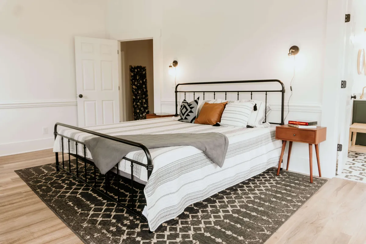 A serene bedroom with patterned bedding atop a comfortable bed, complemented by a nightstand with charming decor and an intricately designed rug. Natural light streams in, enhancing the cozy and peaceful atmosphere.