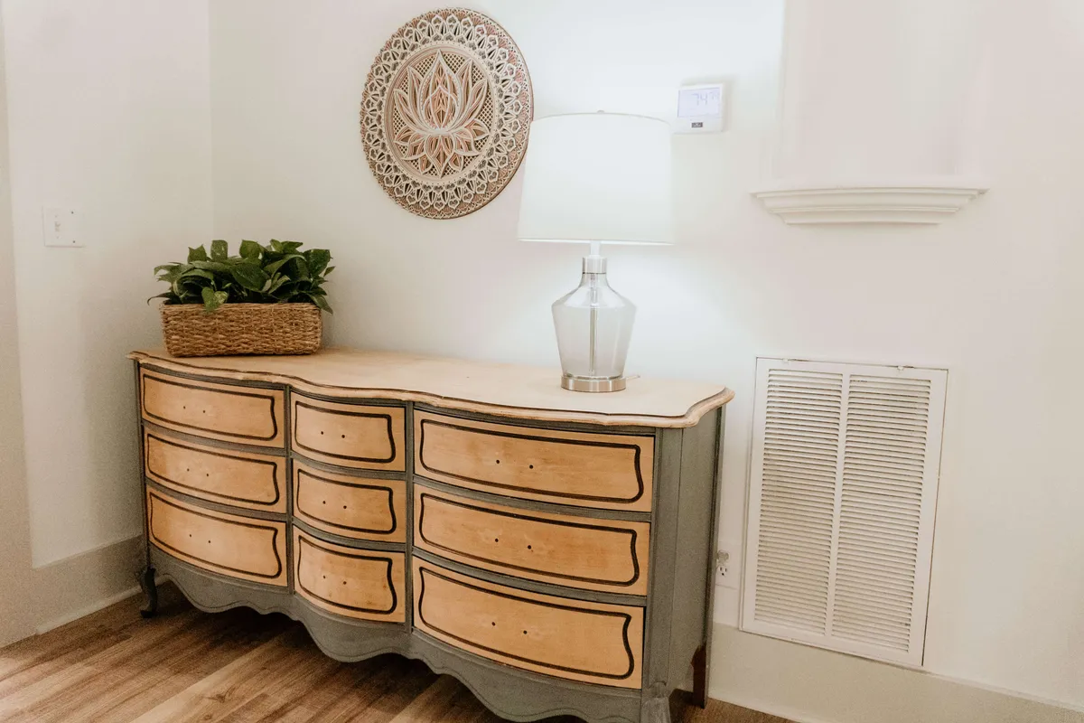 A calm and inviting entryway or sitting area with a well-styled dresser, a glowing lamp, thoughtful wall decor, and a touch of greenery from a potted plant. The scene suggests a warm welcome and a space ideal for intimate conversations or relaxing over morning coffee.